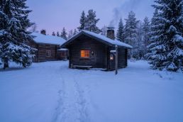 Snow-covered timber sauna cabin beside a farmhouse at dusk, with warm light in the window and smoke from the chimney