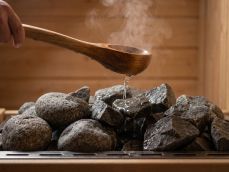 A wooden ladle pours a thin stream of water onto hot dark sauna stones, with a small plume of mist rising