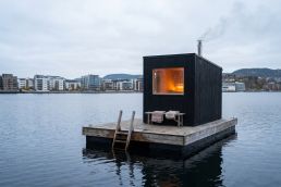 A small dark timber sauna on a wooden pontoon in calm fjord water, with a swim ladder and a waterfront in the distance