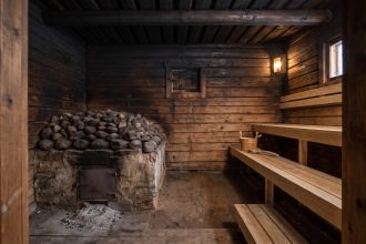 Dark log interior of a smoke sauna with a large stone mound over a masonry base, a closed firing opening and clean wooden benches