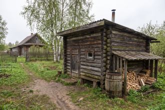 Low log-built banya with a metal chimney, a firewood lean-to and a water barrel in a rural yard