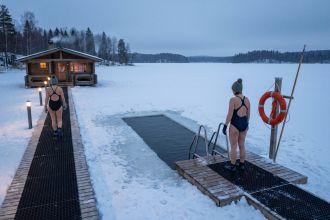 A rectangular hole in snow-covered lake ice with a steel stair and handrails, a lit walkway to a sauna cabin and a lifebuoy post