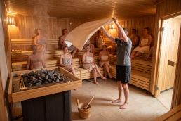 A sauna master swings a large towel overhead in front of the heater while guests sit on three bench tiers